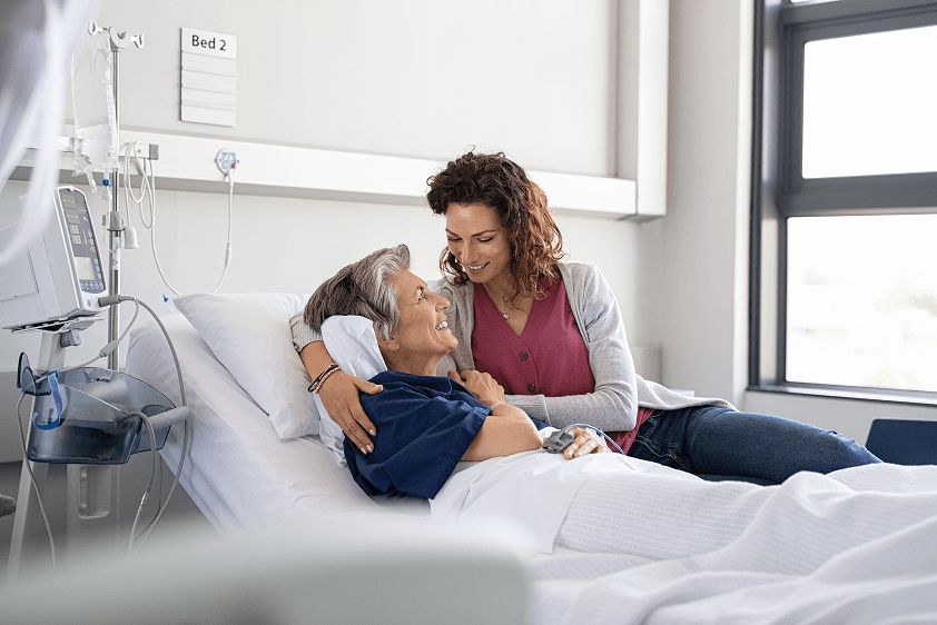 An adult daughter holds her mother's hand by her hospital bedside.